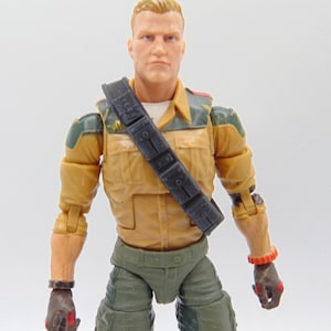 May include: Action figure of a muscular man with short blond hair, wearing a tan military-style jacket, green pants, and a black strap across his chest. The figure has brown gloves and red and orange wristbands. The figure is a collectible toy.