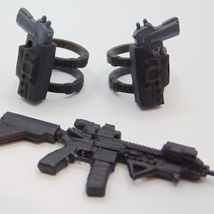 Op de afbeelding: Een set zwarte speelgoedwapens, waaronder een geweer met een vizier en twee pistoolholsters. De holsters zijn bevestigd aan een ringvormige structuur. De items zijn van plastic en staan op een witte achtergrond.