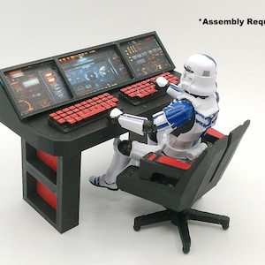 May include: A black desk with red accents and three computer screens, featuring a red keyboard. A white, blue, and black action figure is seated in a black chair at the desk. The text "Assembly Required" is visible.
