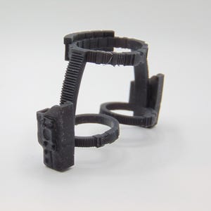 Op de afbeelding: Een donkergrijs, 3D-geprint object met een complex, geometrisch ontwerp. Het heeft meerdere cirkelvormige en rechthoekige componenten die zijn verbonden door geribbelde steunen. Het object lijkt een klein, ingewikkeld mechanisch onderdeel te zijn.