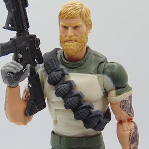 May include: Action figure of a male soldier with a blonde beard and hair, wearing a green and white uniform. The figure is holding a black rifle and has tattoos on the arm. A bandolier of grenades is across the chest.