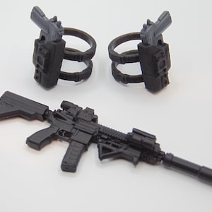 May include: A set of black toy weapons, including a rifle with a scope and silencer, and two pistol holsters attached to circular belt loops. The items are made of plastic and are designed for play or display.