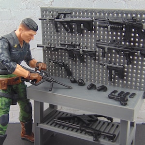 May include: A military action figure with a black uniform and green camouflage pants is arranging black toy weapons on a gray pegboard and table. The weapons include rifles, pistols, and grenades. The scene is set in a military-themed environment.