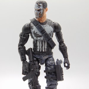 May include: Action figure of a character with a skull face paint design. The figure is wearing a black tactical outfit with a white skull emblem on the chest. It is holding a black rifle and has a holster on its leg.