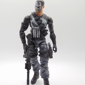May include: A detailed action figure of a character in a black and gray tactical outfit, holding a rifle. The figure has a skull-like face paint design and is posed against a white background. The figure is approximately 6 inches tall.