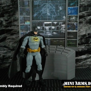 May include: A Batman action figure stands in front of a gray control panel and a wall of computer screens. The figure wears a gray and black suit with a yellow bat symbol and a gold belt. The text "Assembly Required" is visible.
