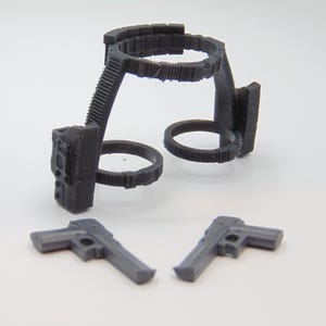 May include: A set of miniature toy weapons, including two gray pistols and a black holster with a belt and two circular holders. The items are made of plastic and are set against a white background.