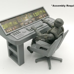 Pode incluir: Uma mesa cinza em miniatura do centro de comando com teclado e várias telas. Uma figura de ação cinza com capacete e colete tático está sentada em uma cadeira cinza, apontando para o console. O texto diz "*Assembly Required."