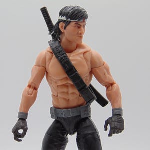 May include: Action figure of a muscular character with a headband, black pants, and gloves. A black strap holds a katana on the figure's back. The figure has a determined expression.