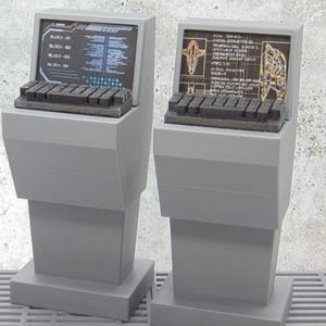 May include: Two gray computer terminal consoles with integrated screens and black keyboards. Each screen displays digital data, including text and technical diagrams. The consoles feature a tapered, upright design with a flat base, presented as a pair.