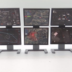 May include: Three sets of dual-stacked computer monitors on gray stands. Each screen displays digital interfaces, including topographical maps, data charts, and tactical grid overlays with various icons and text elements in a dark color scheme.