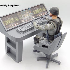 Könnte beinhalten: Ein grauer Kommandozentrale-Schreibtisch mit detailliertem Bildschirm und Tastatur, begleitet von einer sitzenden Actionfigur. Der Schreibtisch hat ein Design mit zwei Regalen und den Text "*Assembly Required". Die Figur trägt eine graue Uniform und eine schwarze Mütze.