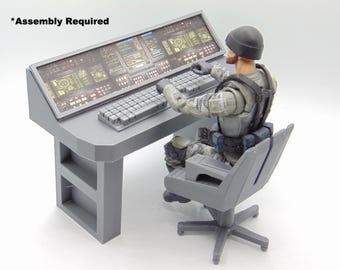 Op de afbeelding: Een grijze commandocentrumtafel met een gedetailleerd scherm en toetsenbord, vergezeld van een zittend actiefiguur. De tafel heeft een ontwerp met twee planken en de tekst "*Assembly Required". Het figuur draagt een grijs uniform en een zwarte muts.