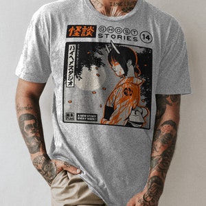 Op de afbeelding: Een grijs T-shirt met een grafische print van een comicboekomslag met een vrouw met hoorns en een rood-oranje outfit. De tekst op de omslag luidt "怪談 GHOST STORIES 14" en "バイヘイスタジオ PA-HEME STUDIO".