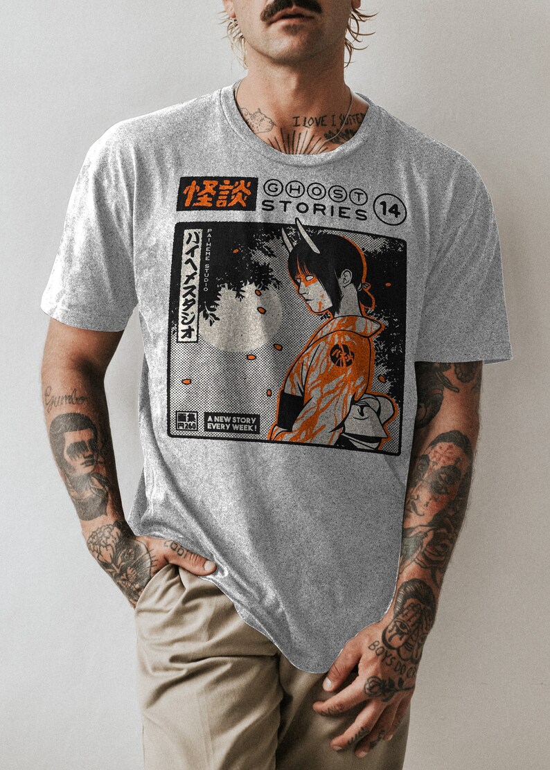 Op de afbeelding: Een grijs T-shirt met een grafische print van een comicboekomslag met een vrouw met hoorns en een rood-oranje outfit. De tekst op de omslag luidt "怪談 GHOST STORIES 14" en "バイヘイスタジオ PA-HEME STUDIO".