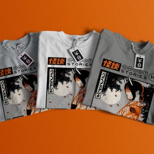 Op de afbeelding: Drie grijze, witte en lichtgrijze T-shirts met een grafische print van een vrouw met hoorns en Japanse tekst die "怪談" leest, wat "Spookverhalen" betekent.
