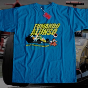 Puede incluir: Una camiseta azul brillante con una imagen de dibujos animados de un piloto de carreras y el texto "FUMANDO ALONSO GRAN FUMADA DE EUROPA".