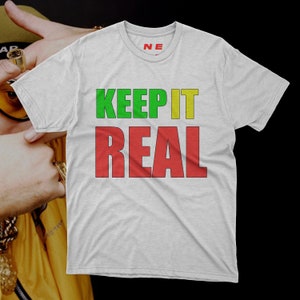 Puede incluir: Una camiseta blanca con las palabras "KEEP IT REAL" impresas en letras verdes, amarillas y rojas.