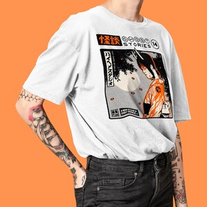 Op de afbeelding: Wit T-shirt met een zwarte en oranje grafische print van een manga-achtig personage met hoorns. De grafiek bevat Japanse tekst en de woorden "Ghost Stories" en "A New Story Every Week".