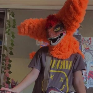 May include: A person wearing an orange furry dinosaur costume with a red mohawk and a black t-shirt with a yellow smiley face graphic and the word "Nirvana" printed on it. The person is holding a brown and black electric guitar.