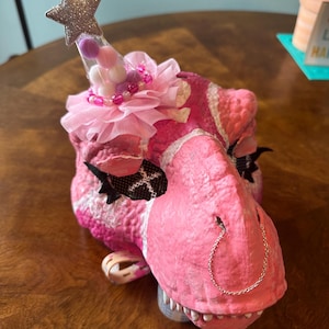 May include: Pink dinosaur mask with a white and pink pattern. The mask has a silver star on top with pink and white pom poms and beads. A silver chain hangs from the nose.