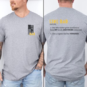 Może przedstawiać: Szary T-shirt z czarno-ż&oacute;łtym wzorem amerykańskiej flagi z przodu i tekstem "Line Dad [NOUN] 1. One who makes great sacrifices to keep MY family AND YOURS connected. 2. Like a regular dad but TOUGHER." z tyłu.