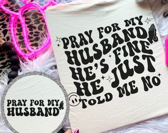 May include: A cream-colored t-shirt with the text "PRAY FOR MY HUSBAND HE'S FINE HE JUST TOLD ME NO" in black, retro-style lettering. A matching round pocket accent is also visible.
