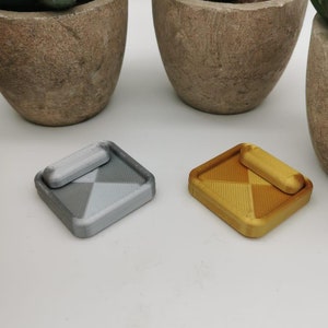 Könnte beinhalten: Zwei kleine, quadratische Aschenbecher mit geometrischem Design. Einer ist silberfarben und der andere goldfarben. Jeder Aschenbecher hat eine kleine, rechteckige Ablage. Die Aschenbecher werden auf einer weißen Oberfläche mit Topfpflanzen im Hintergrund präsentiert.
