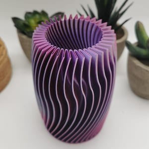 Puede incluir: Un jarrón de color morado y rosa impreso en 3D con un diseño ondulado y acanalado. El jarrón está hecho de plástico y tiene forma cilíndrica.