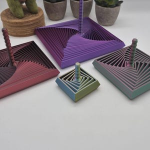 Peut inclure: Quatre toupies carrées avec un design géométrique en spirale à plusieurs niveaux. Les toupies présentent des transitions de couleurs dégradées, notamment violet, rose, bleu sarcelle et vert. Chaque toupie a une poignée centrale striée.