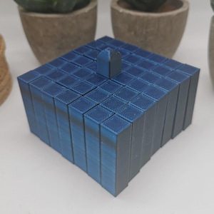Può includere: Una scatola blu a forma di cubo fatta di piccoli blocchi impilati. La scatola ha un piccolo pezzo rettangolare blu in cima.