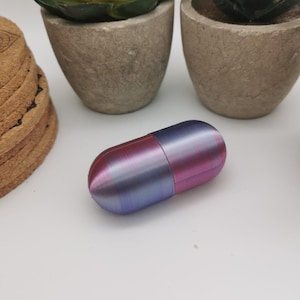 Op de afbeelding: Een kleine, pilvormige container met een metallic afwerking. De ene helft is lichtblauw met een subtiele glans, terwijl de andere helft donkerroze is. De container ligt op een wit oppervlak.