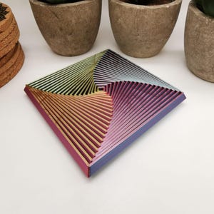Könnte beinhalten: Ein quadratisches, mehrfarbiges Dekotablett mit geometrischem Design. Das Tablett weist einen Farbverlauf auf, darunter Rosa, Lila, Gelb und Blau. Das Tablett besteht aus geschichtetem Material und hat eine zentrale quadratische Vertiefung. Es ist ein dekorativer Artikel.