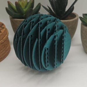 Könnte beinhalten: Ein teal-farbenes, kugelförmiges, lasergeschnittenes Dekorationsobjekt mit geschlitztem Design. Das Objekt befindet sich vor mehreren Topf-Sukkulenten und einem kleinen dekorativen Holzgegenstand.