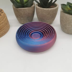 May include: A circular soap dish with a gradient of blue and pink hues. The dish features concentric circles. The soap dish is made of a 3D printed material. The background includes small potted plants.