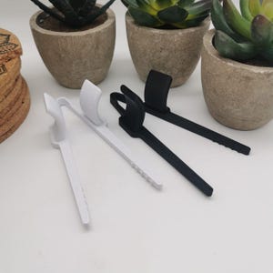 Op de afbeelding: Twee paar plastic clips, een zwarte en een witte, ontworpen om items bij elkaar te houden. De clips zijn gemaakt van flexibel materiaal en hebben een eenvoudig ontwerp.