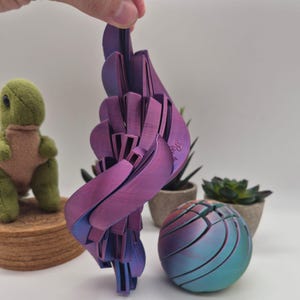 Op de afbeelding: Een 3D-geprinte gearticuleerde sculptuur met een verloop van paarse en blauwe kleuren met een metallic afwerking. Het object heeft een complexe, gelaagde en gesegmenteerde structuur met gebogen, vleugelachtige panelen die verbonden zijn voor beweging.