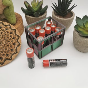 以下が含まれることがあります： オレンジと黒の単三電池が詰まった緑色のプラスチック製の箱。箱は、茶色のコースターと緑色の植物が入った鉢のあるテーブルの上に置かれています。