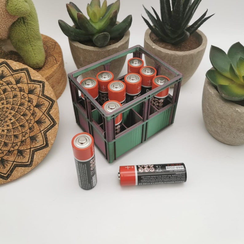 以下が含まれることがあります： オレンジと黒の単三電池が詰まった緑色のプラスチック製の箱。箱は、茶色のコースターと緑色の植物が入った鉢のあるテーブルの上に置かれています。