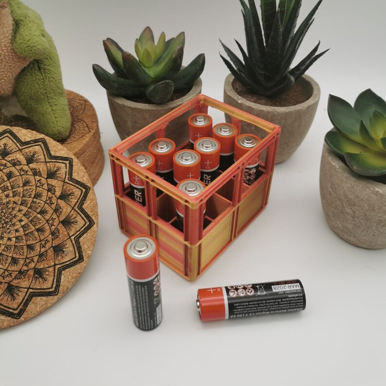 以下が含まれることがあります： オレンジと白のプラスチック製の電池ホルダーに、単三電池が10本入っています。ホルダーは、コルクコースターと3つの鉢植えの植物のあるテーブルの上に置かれています。