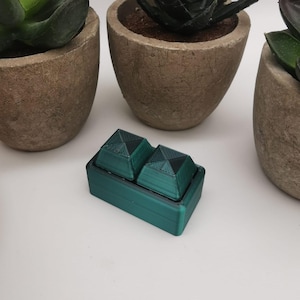 Könnte beinhalten: Eine kleine, tealgrüne Box mit zwei pyramidenförmigen Deckeln. Die Deckel sind aus dunkelgrünem Kunststoff und haben eine strukturierte Oberfläche.