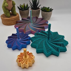 Puede incluir: Cuatro peonzas impresas en 3D de diferentes tamaños y colores. La más grande tiene un degradado multicolor, seguida de una verde azulada, una azul y una pequeña amarilla. Cada peonza tiene un mango vertical central y una base estrellada.
