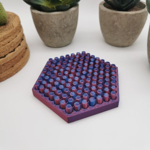 May include: A hexagonal coaster with a textured surface. The surface is covered in small, raised, cylindrical shapes in alternating shades of blue and pink.