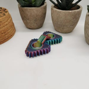 May include: Two iridescent, gear-shaped fidget toys with a rainbow finish. The gears have a central ring and a handle connecting them. The toys are on a white surface, with potted plants in the background.