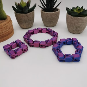 Può includere: Tre braccialetti flessibili realizzati con pezzi di plastica collegati. I braccialetti sono di dimensioni diverse e presentano un gradiente di viola e blu.