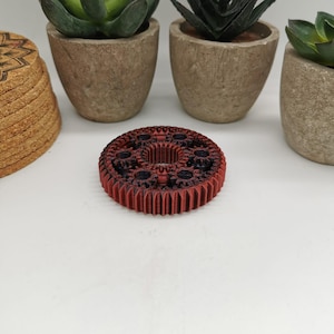 Pode incluir: Um objeto em forma de engrenagem vermelho e preto com detalhes intrincados. A engrenagem tem um design circular com várias engrenagens menores no centro. O objeto está sobre uma superfície branca, com pequenas plantas em vasos no fundo. Diâmetro: aprox. 7 cm.