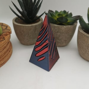 Pode incluir: Uma escultura piramidal geométrica com um gradiente de vermelho e azul marinho. A pirâmide tem uma série de recortes curvos em forma de onda. A escultura é exibida em frente a várias suculentas em vasos.