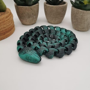 Pode incluir: Um colar em forma de cobra verde e preto feito de contas interligadas. O colar está enrolado em espiral com a cabeça da cobra na frente.