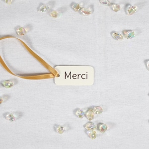 Könnte beinhalten: Ein weißes Etikett mit dem Wort "Merci" in schwarzer Schrift, befestigt an einem goldenen Band, liegt auf einer weißen Oberfläche. Iridescent schimmernde, kristallartige Verzierungen sind verstreut.