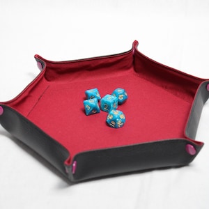 May include: A black and red dice tray with a square shape and rounded corners. The interior is red, and the exterior is black with pink accents. Five blue dice with gold numbers are inside the tray. This tray is designed for tabletop gaming.